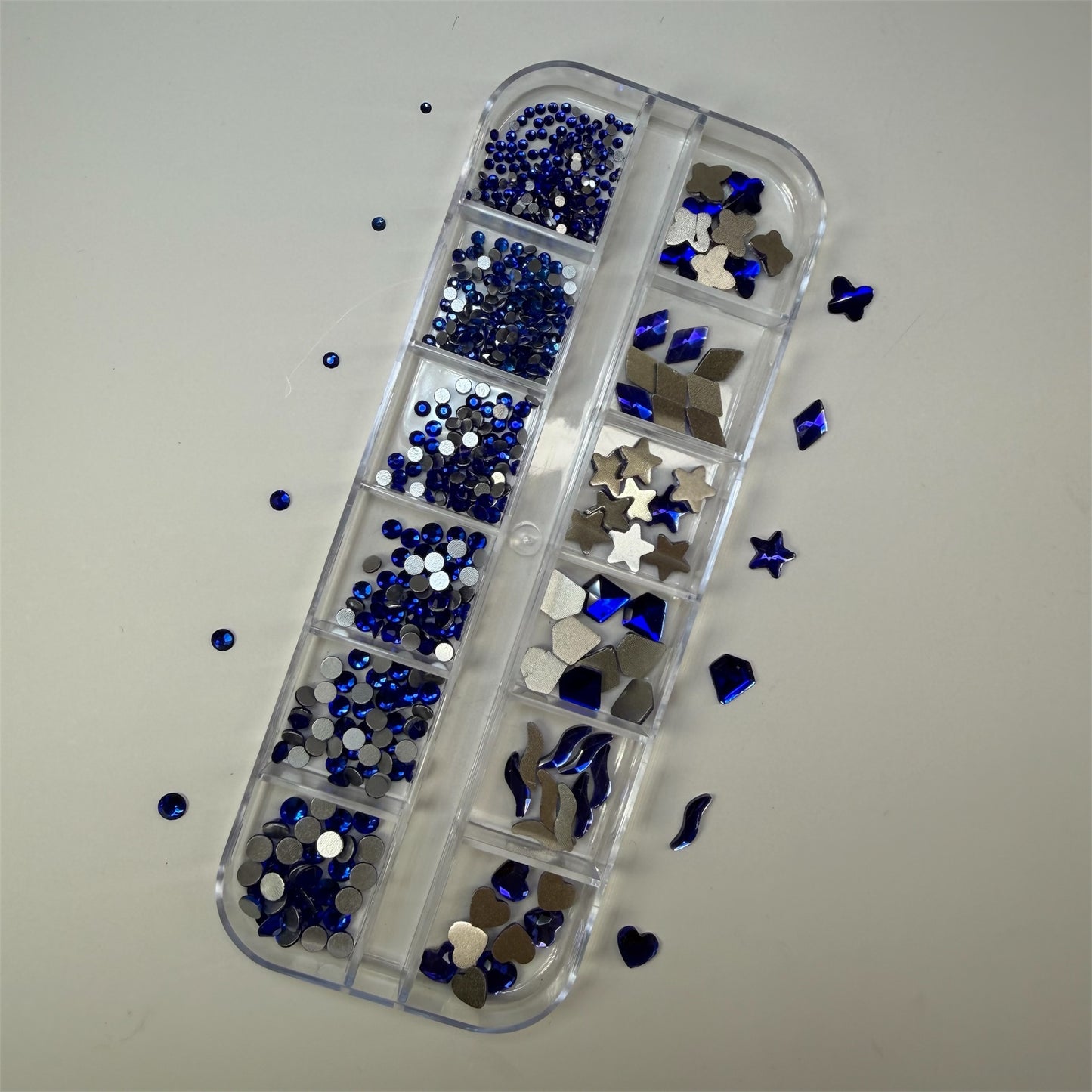 Blue mixed nail rhinestones and flatback charms in assorted shapes and sizes, including hearts, stars, diamonds, and rounds, organized in a clear plastic nail art storage case.